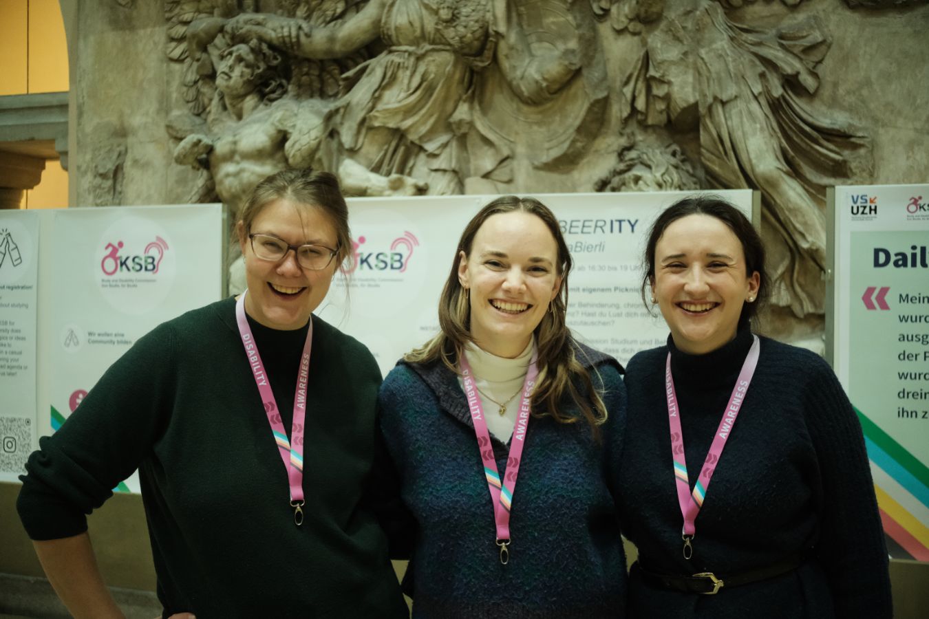 Plakatausstellung: Drei Mitglieder der KSB-VSUZH stehen nebeneinander und lächeln vor Plakaten zum Thema Disability Awareness im Lichthof der UZH.